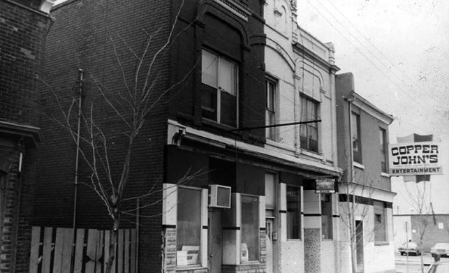 a black and white photo of a street corner and one of the building having the sign copper johns entertainment